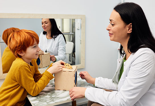 A clinician in a white coat sits at a small table with a child who has bright red hair and is playing with a wooden toy.