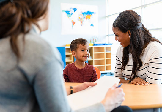 An elementary-aged boy looks at a woman sitting next to him at a table, while another adult looks on in the foreground.