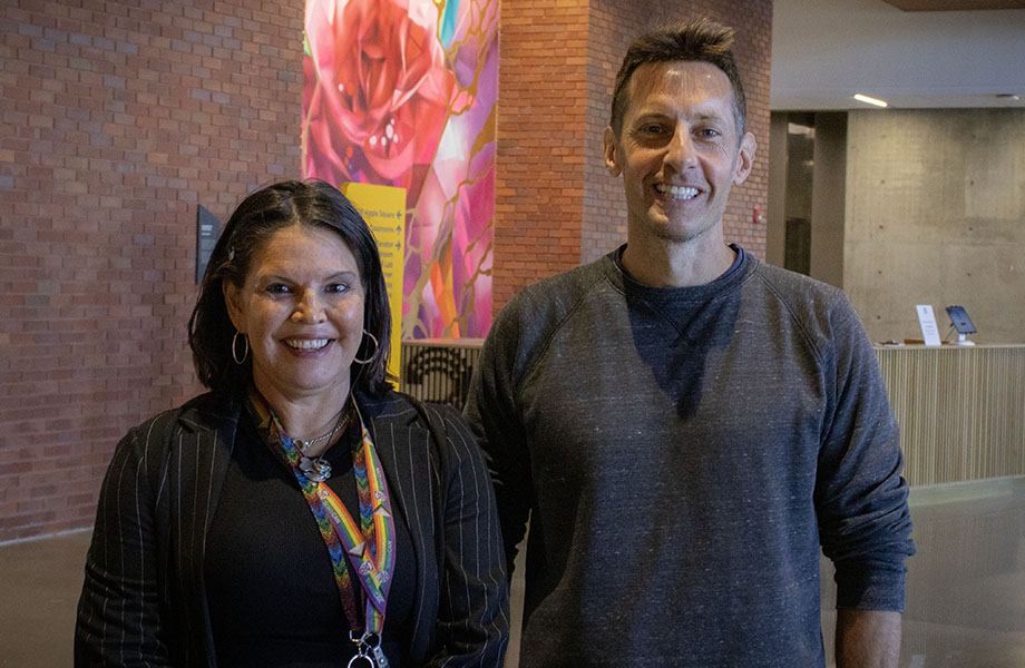 Two adults stand in a building with a brick wall and artwork 