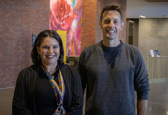 Two adults stand inside a building with a pink flower design artwork behind them. 