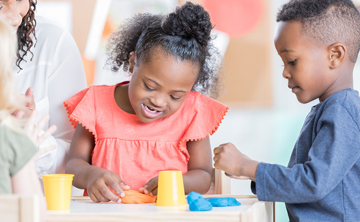 Two young children play with a colorful dough while an adult looks on.