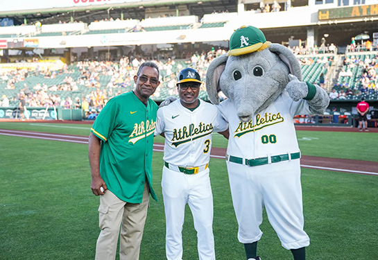 Two men in baseball jerseys standing next to costumed character.