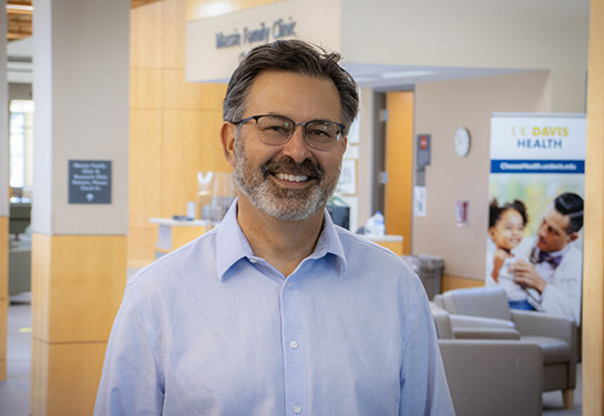 A man with brownish gray hair, wearing a light blue button-down shirt, stands inside a medical building at UC Davis Health. 