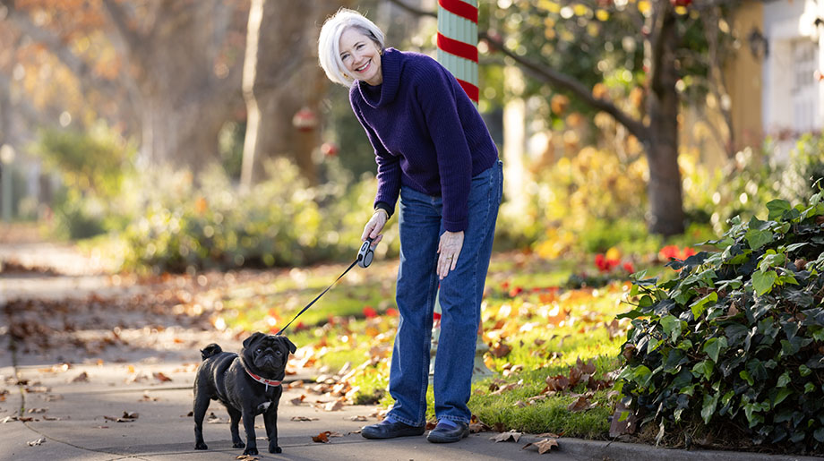 A woman with white hair looks down at a black Pug whom she has on a leash on a tree-lined street.