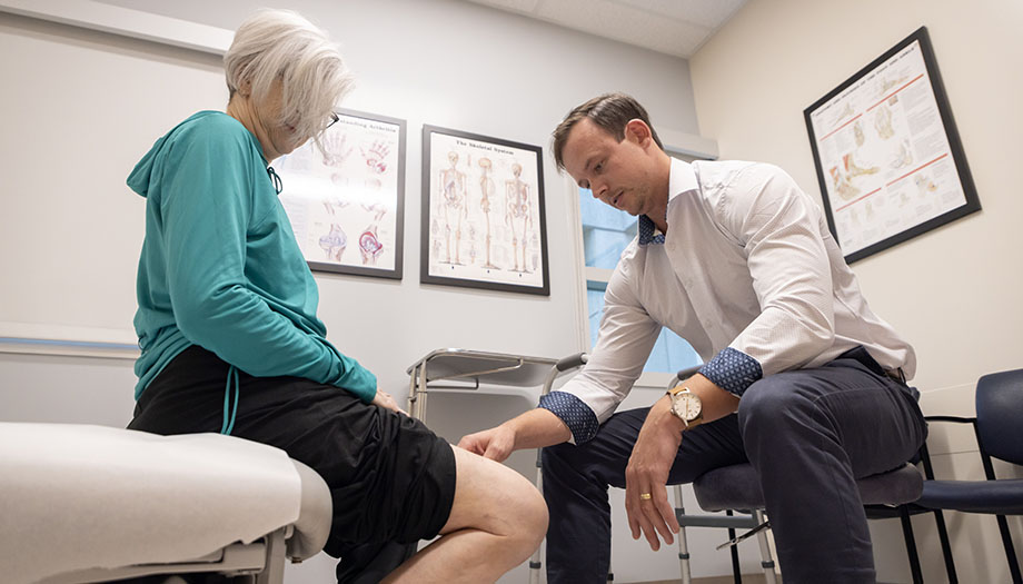 A woman sits on an exam table while a physician in a white coat examines her knee.
