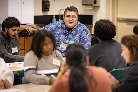 Man with dark hair and blue and white shirt talking with students at desk.