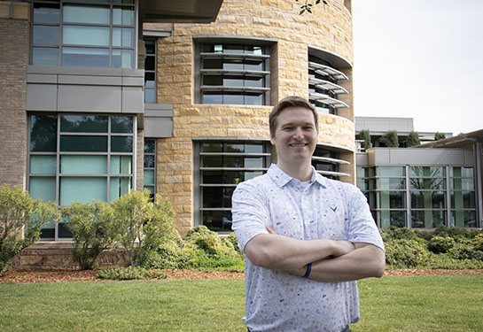 A man wearing a lavender shirt, with his arms crossed, stands outside in front of the UC Davis MIND Institute