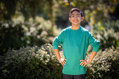 Dark haired boy with green shirt and hands on hips standing outside