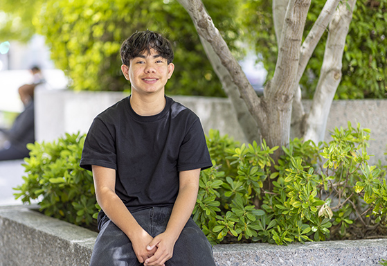 Teen boy in black shirt sitting outside by a tree.