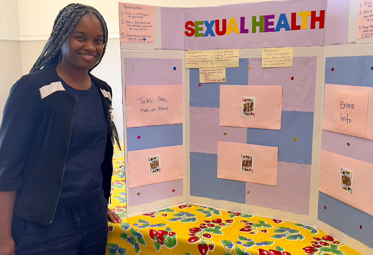 Nursing student in blue dress stands in front of colorful poster board presentation with title that reads &ldquo;sexual health&rdquo;