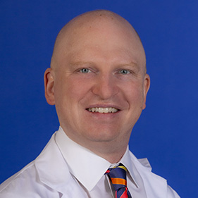 Headshot of smiling bald man in a white doctors coat, white shirt and red and blue tie.