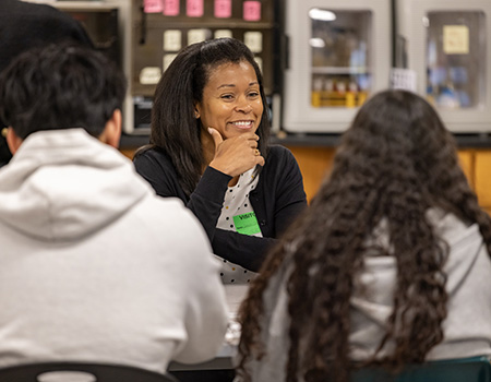 Woman smiles at students surrounding her at desk.