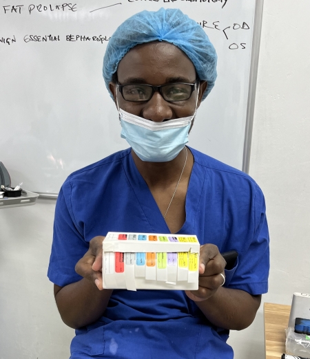 A doctor wearing black glasses and blue scrubs holds a box with colorful printed labels. 