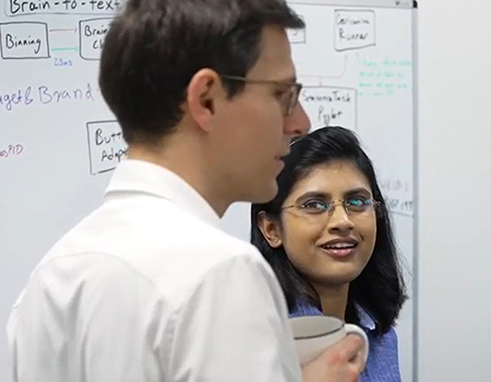 Sergey Stavisky in a white shirt and eyeglasses speaks with Maitreyee Wairagkar in front of a whiteboard with writings.