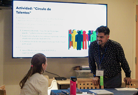 A woman, seated, talks to a man, standing, in front of a whiteboard containing a presentation in Spanish. 