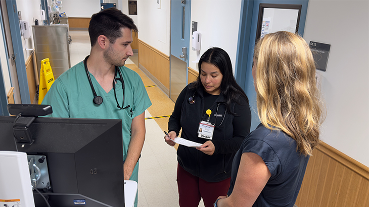 Three physicians discuss notes about patients in a hospital hallway in front of a computer workstation.