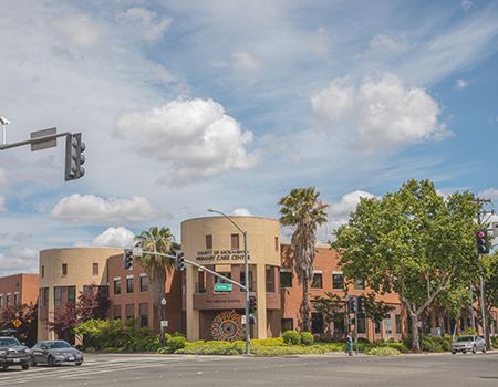 Building with sign that states County of Sacramento Primary Care Center at a sunny intersection with cars and palm trees.