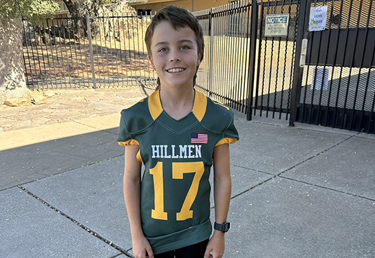 Smiling boy in a green and gold football jersey with a orthopaedic boot on one leg and a white tennis shoe on the other.
